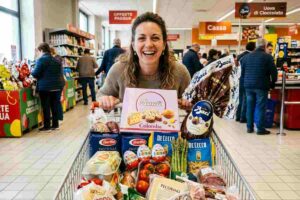 Donna sorridente con carrello pieno per la spesa di Pasqua 2026 tra le corsie del supermercato con uova di cioccolato e colombe.