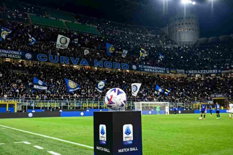 Stadio San Siro gremito per Inter-Roma, pallone ufficiale Serie A su colonnina a bordo campo e tifosi nerazzurri in Curva Nord di sera.